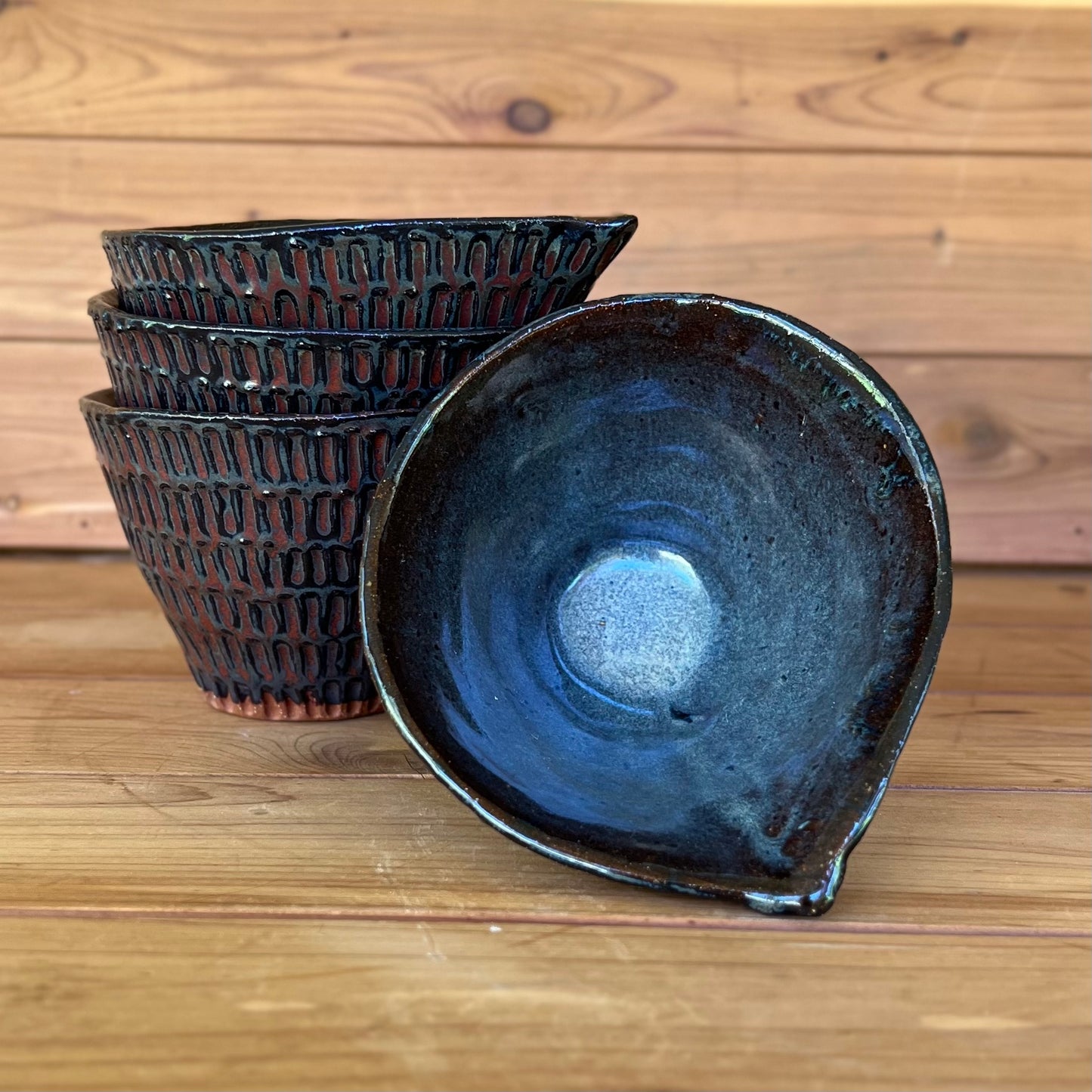 four little carved ceramic espresso cups against a wood background with a black and red bronze glaze