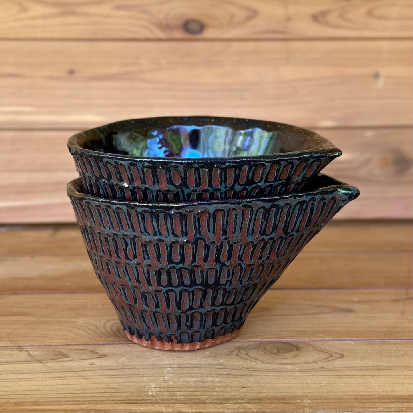 stack of two little carved ceramic espresso cups against a wood background with a black and red bronze glaze
