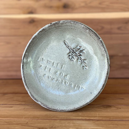 image of small handmade black clay plate glazed with a silver cream glaze and with a botanical stamp and the saying "It will all end eventually" stamped into the clay