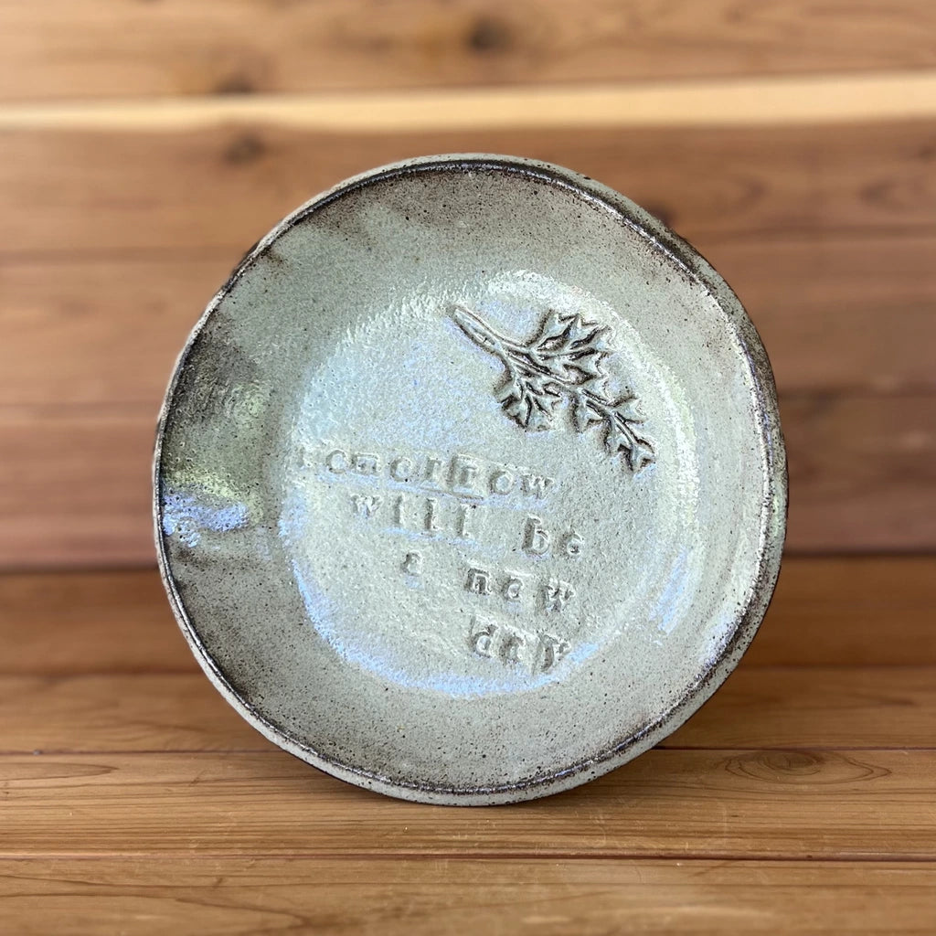 image of small handmade black clay plate glazed with a silver cream glaze and with a botanical stamp and the saying "tomorrow will be a new day" stamped into the clay